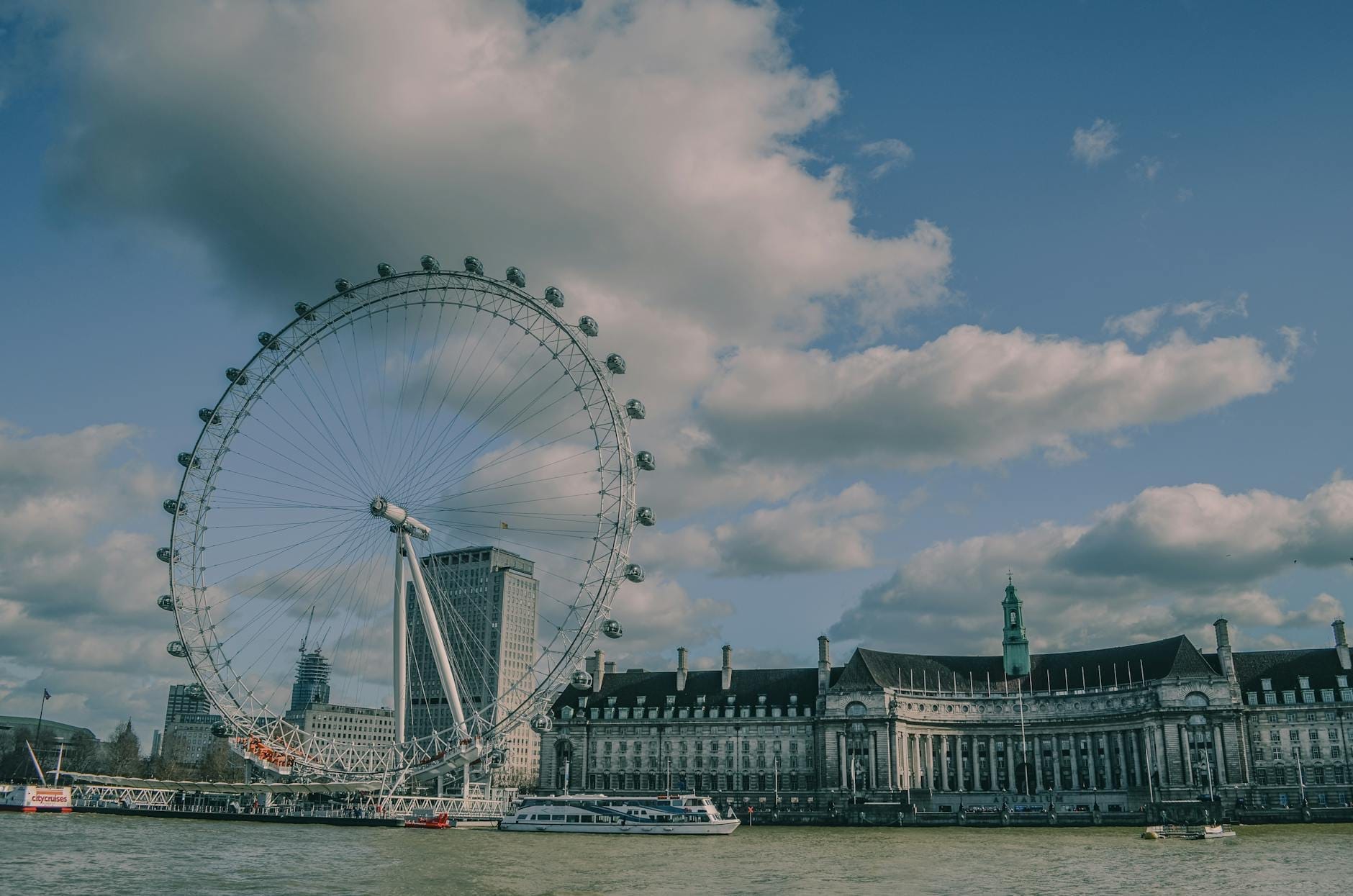 La ruota panoramica del London Eye si staglia sul Tamigi, con County Hall e le barche sul fiume sotto un cielo azzurro con nuvole bianche e soffici.