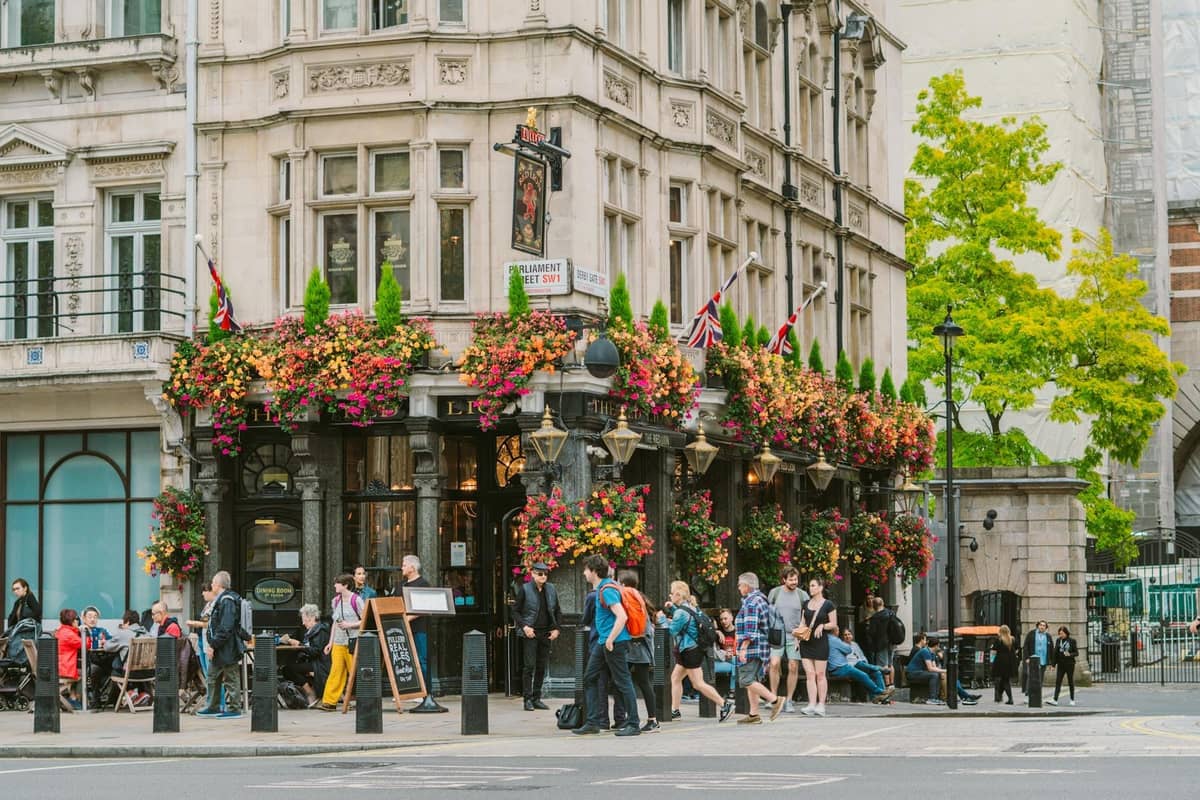 Um clássico pub londrino com arquitetura ornamentada, cestas de flores coloridas e pessoas reunidas do lado de fora desfrutando da atmosfera animada.