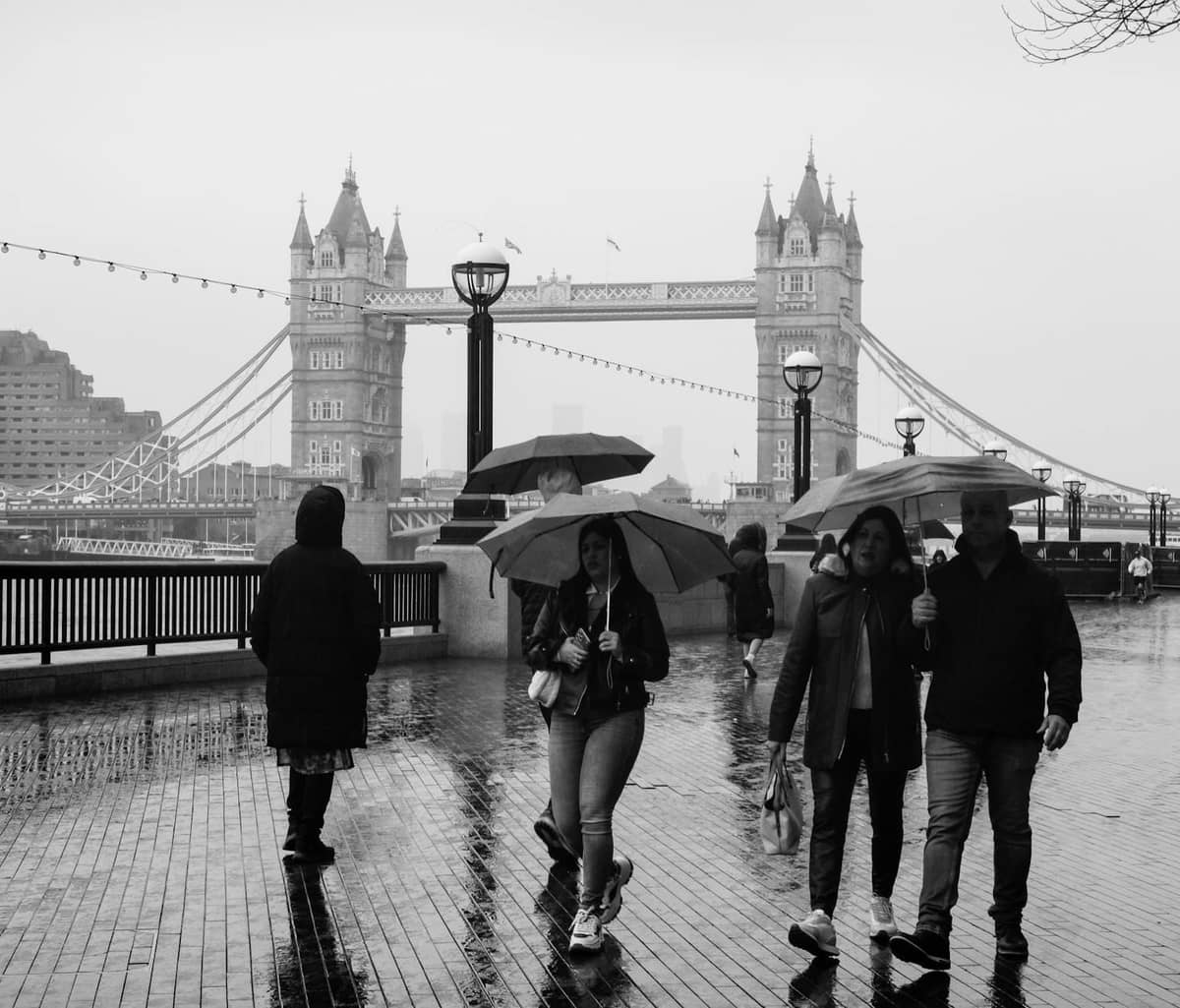 Pessoas caminhando com guarda-chuvas em um dia chuvoso perto da Tower Bridge em Londres, calçada molhada e céu nublado.