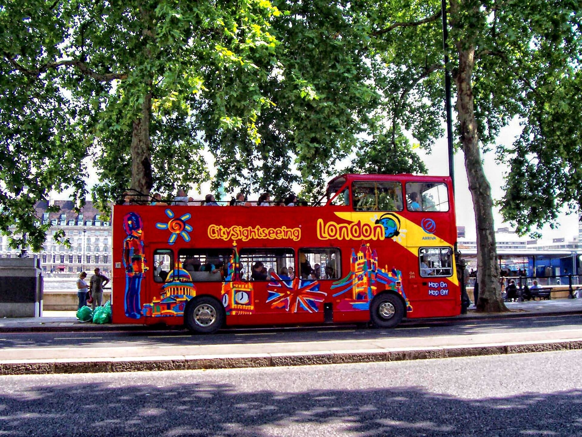 Ônibus de dois andares vermelho brilhante City Sightseeing London hop-on hop-off estacionado sob árvores frondosas, com marcos icônicos da cidade e o rio ao fundo.