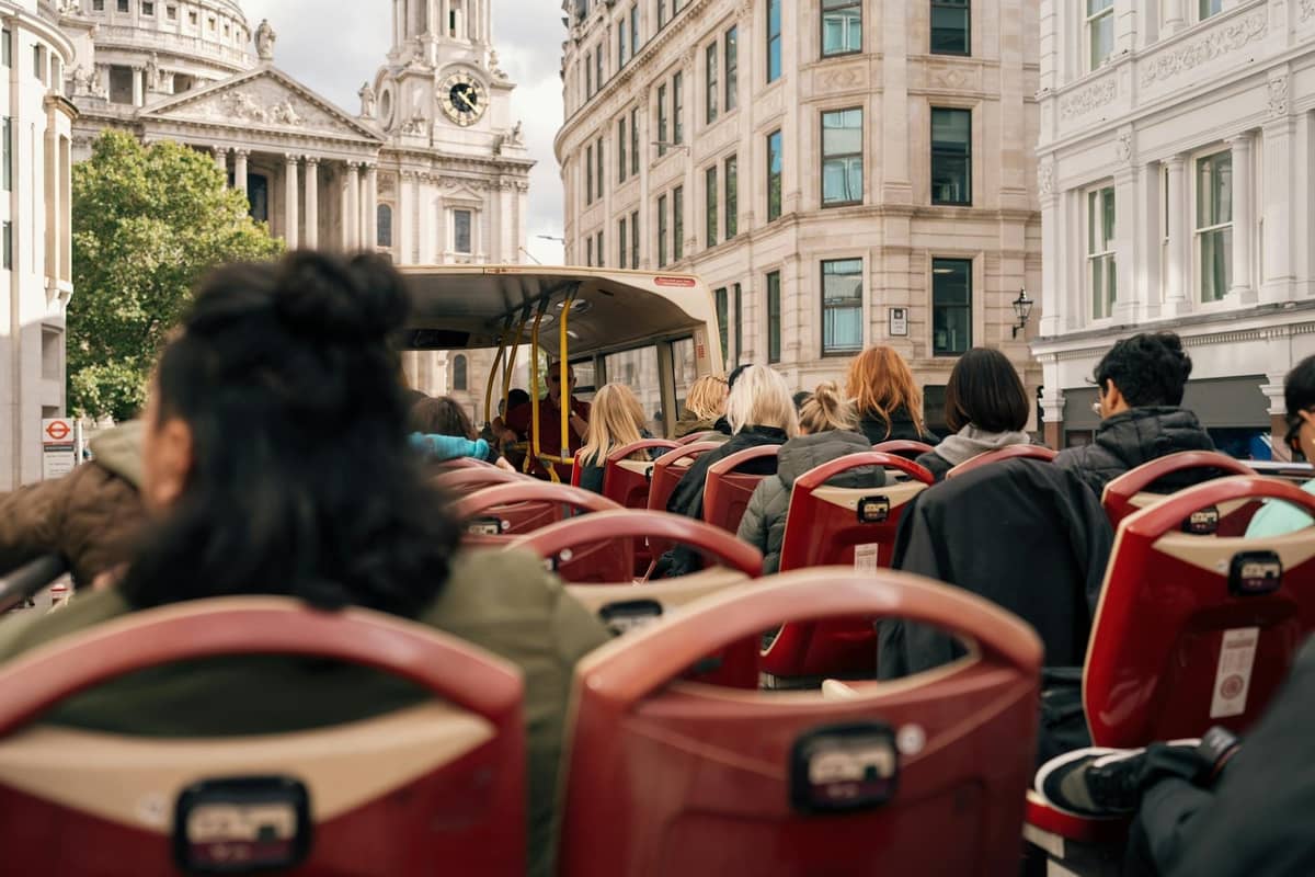 Passageiros sentados no deck superior aberto de um ônibus vermelho hop-on hop-off de Londres, passando por edifícios históricos de pedra e a Catedral de St. Paul's.