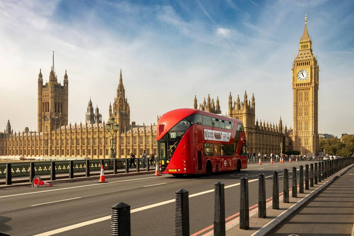 Um ônibus vermelho de dois andares de Londres passa em frente ao Big Ben e ao Houses of Parliament sob um céu limpo, mostrando uma vista clássica de rota turística.