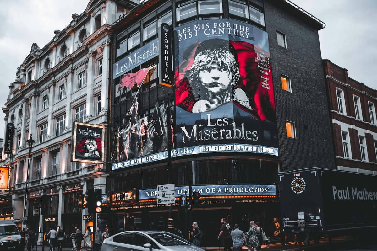 Fachada do Sondheim Theatre de Londres ao entardecer, bem iluminada com grandes letreiros de Les Misérables e pessoas caminhando do lado de fora