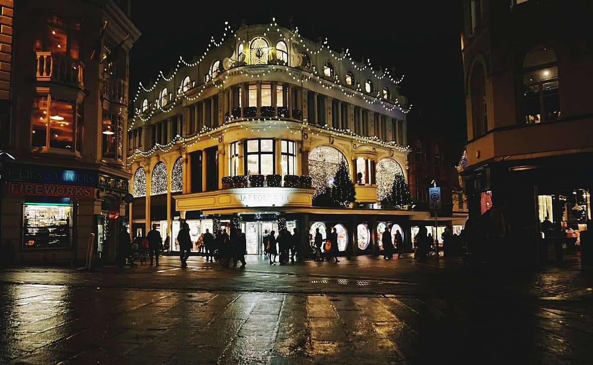 Uma rua de Londres bem iluminada à noite em dezembro, com um grande edifício decorado com luzes festivas e ornamentos natalinos, o pavimento molhado refletindo o brilho.