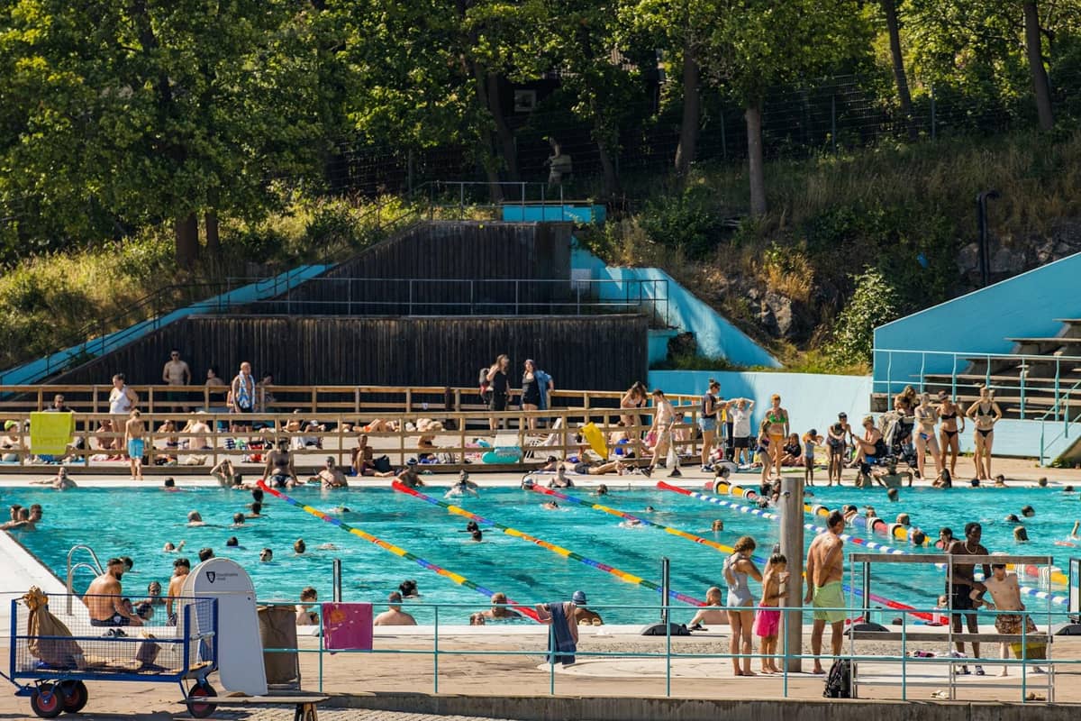 Uma movimentada piscina pública ao ar livre com multidões de pessoas aproveitando o sol, rodeada de árvores e paredes azuis, em um dia de verão.