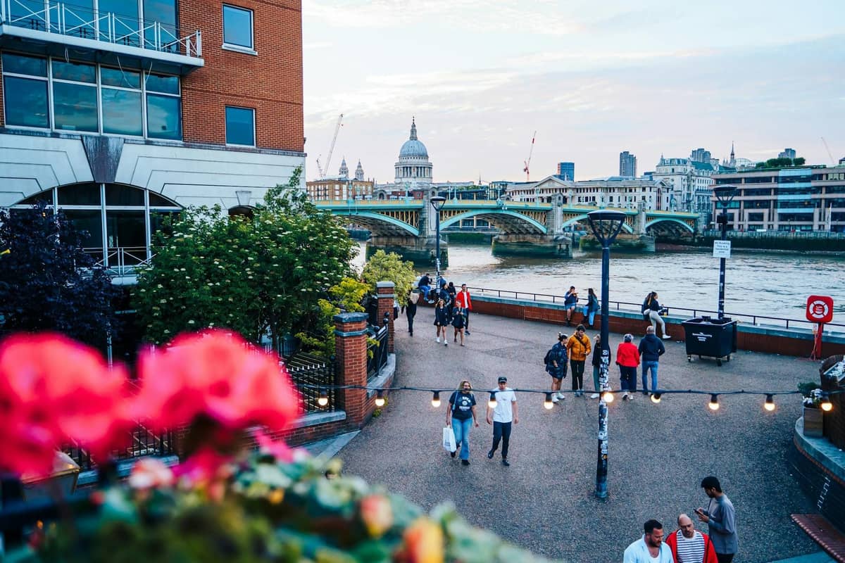 Pessoas passeando pelo calçadão à beira do Tâmisa em Londres, com a Catedral de St. Paul's e uma ponte visíveis ao fundo.
