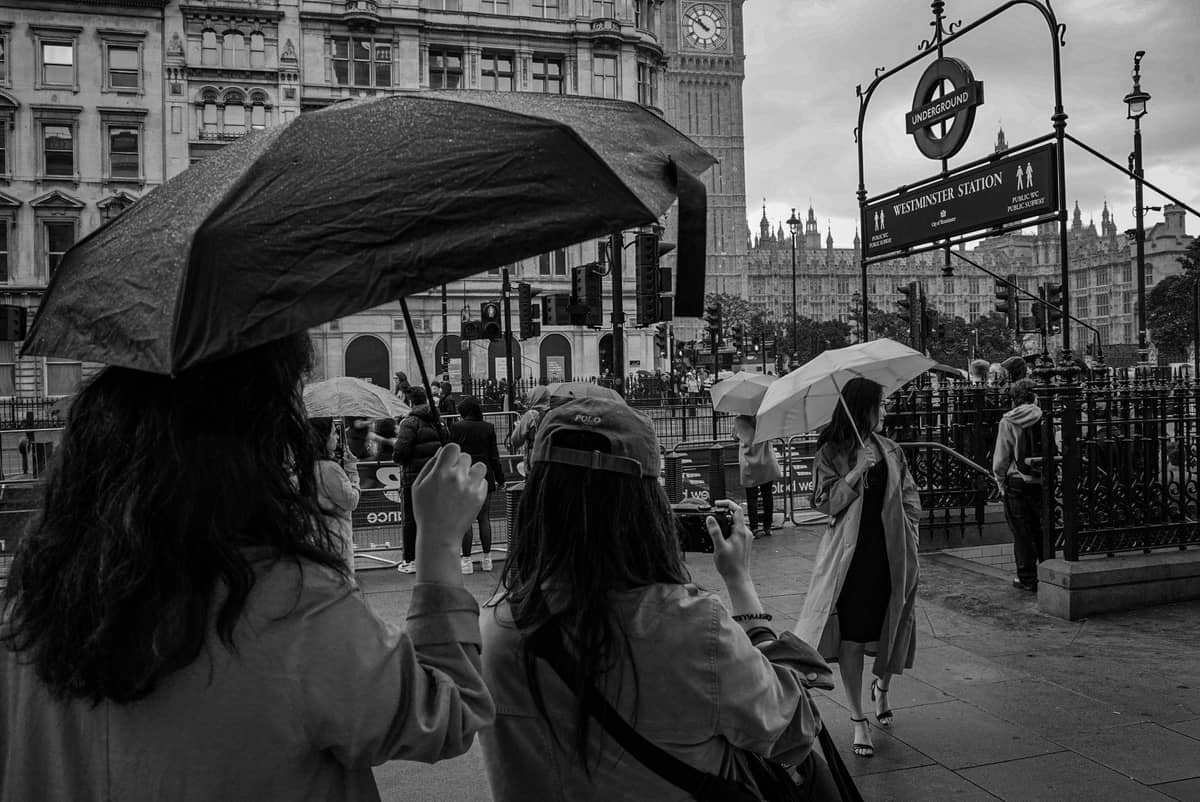 Pessoas com guarda-chuvas em frente à estação de Westminster e ao Big Ben na chuva, mostrando o clima típico de verão em Londres.