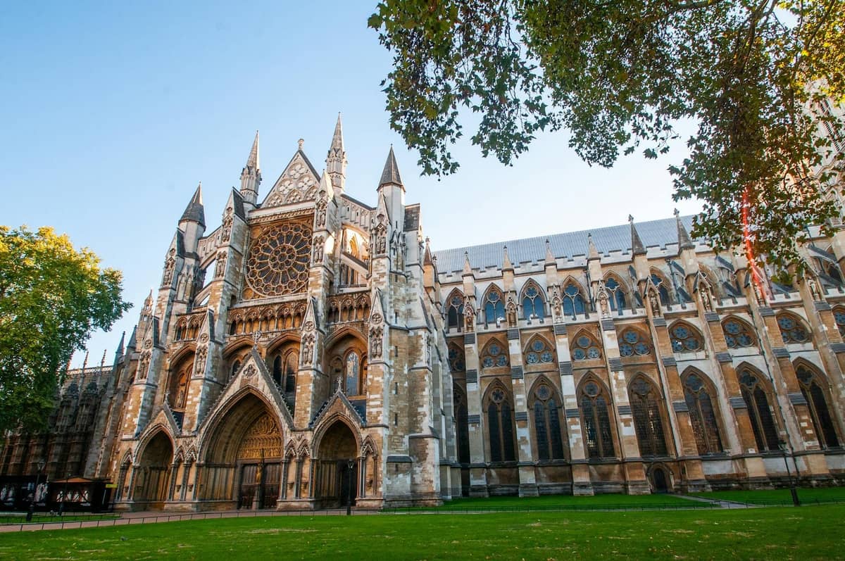 Vista em grande angular da fachada principal e do jardim da Westminster Abbey, com arcos góticos, grande vitral em rosácea e árvores enquadrando o fundo.