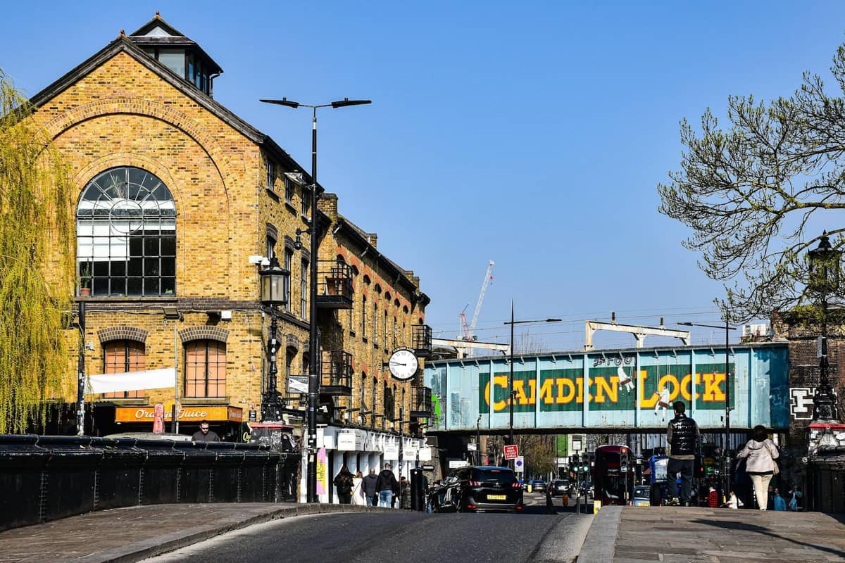 Vista de Camden Lock em Londres com a icônica ponte ferroviária azul e amarela de Camden Lock, edifícios do mercado e pessoas caminhando nas proximidades.