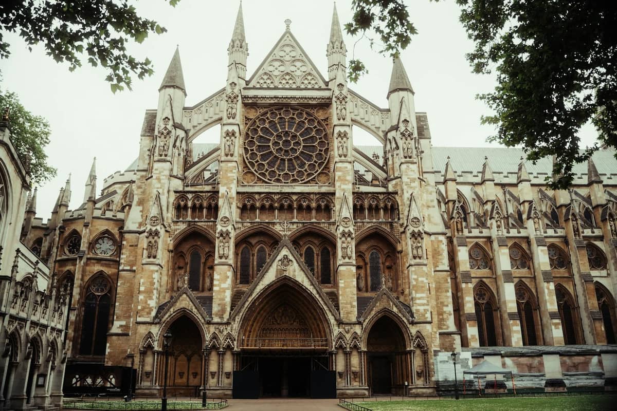 Vista frontal completa do exterior da Westminster Abbey com intrincada obra em pedra, janela em rosa e torres pontiagudas, emoldurada por árvores frondosas em ambos os lados.