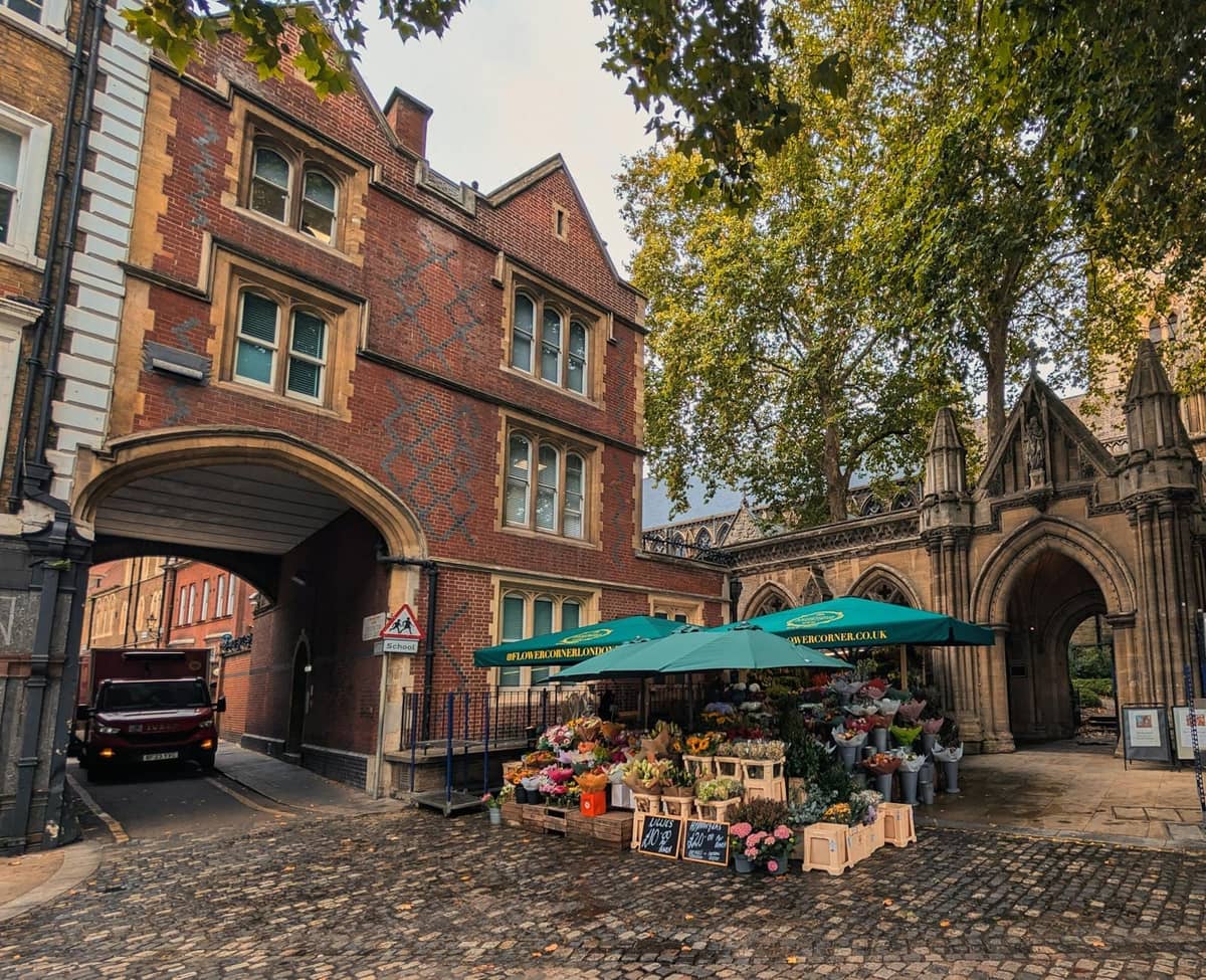 Banca de mercado de flores sob guarda-sóis verdes em frente a históricos edifícios de tijolo vermelho e um arco de pedra numa rua de paralelepípedos em Londres.