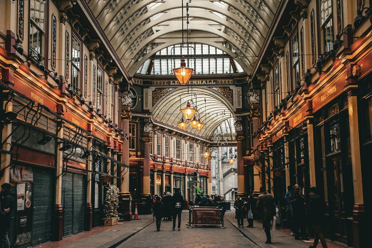 Vista ampla do interior ornamentado com teto de vidro do histórico Leadenhall Market de Londres, com pessoas caminhando sob a arquitetura vitoriana e luzes quentes.
