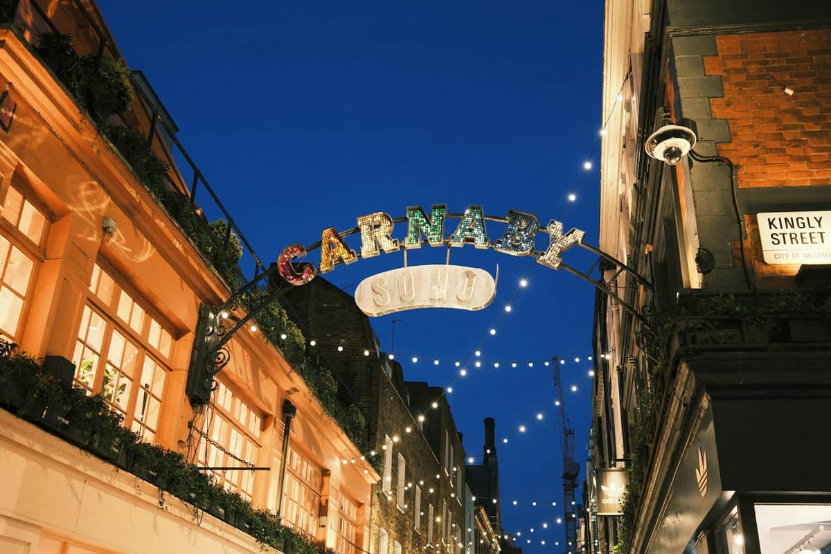 Entrada da Carnaby Street iluminada à noite com luzes festivas, rodeada por icônicos edifícios de Londres, capturando uma famosa área de vida noturna.