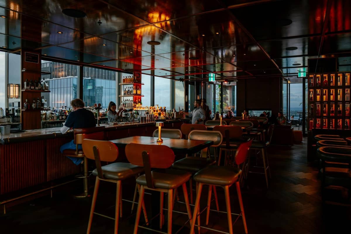 Interior de bar moderno e estiloso ao pôr do sol com vistas para a cidade através de grandes janelas, pessoas sentadas no balcão e nas mesas, e prateleiras bem abastecidas.