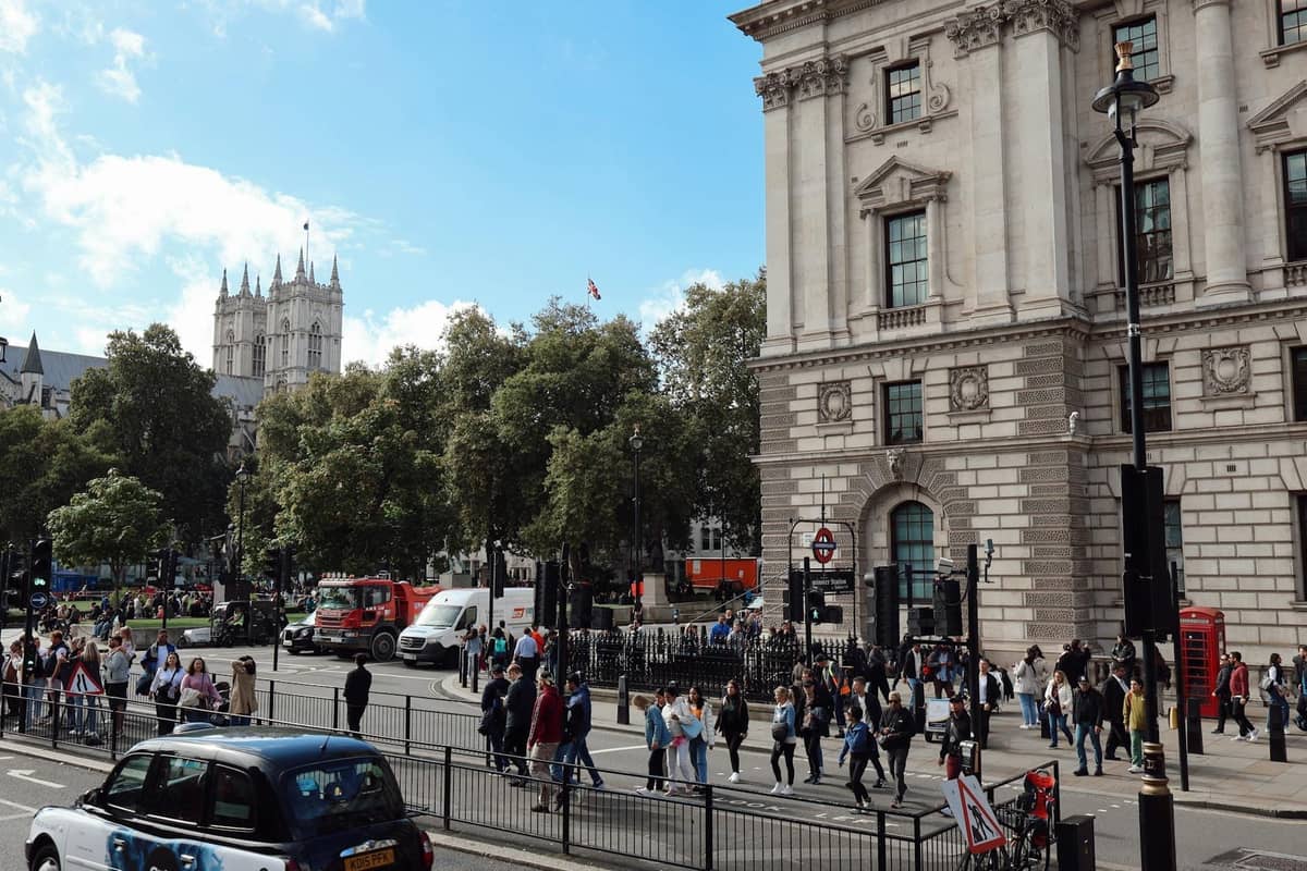 Movimentada cena de rua em Londres com Westminster Abbey ao fundo, pessoas caminhando, tráfego e edifícios clássicos da cidade.