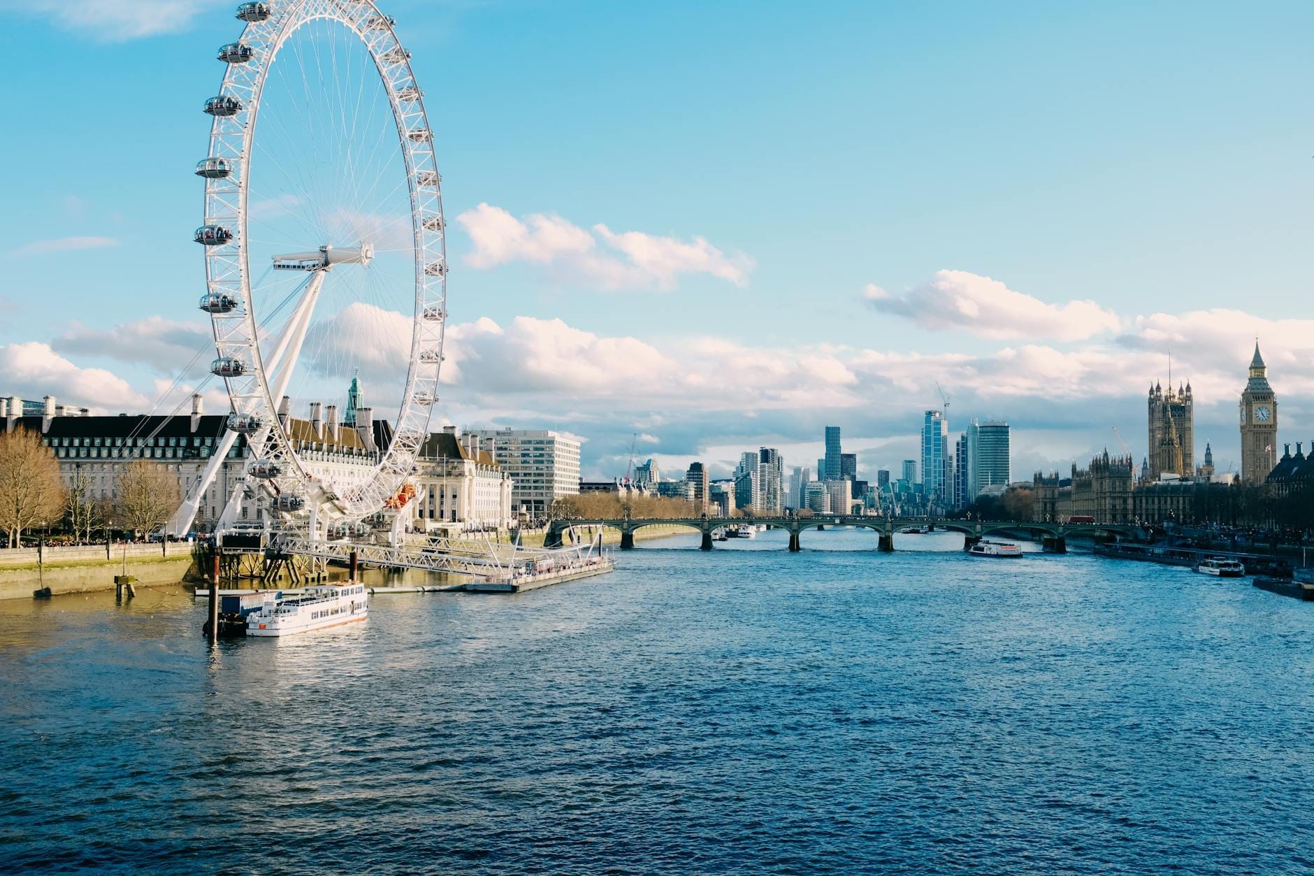 O London Eye, o Rio Tâmisa e o Big Ben sob um céu azul limpo com barcos e edifícios da cidade visíveis ao longo da água.