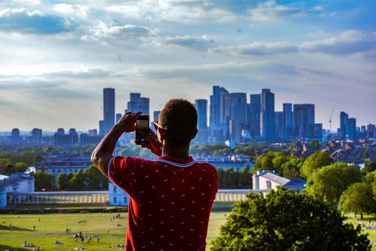 Pessoa fotografando o skyline de Londres a partir de Greenwich Park num dia ensolarado, com marcos da cidade e gramados verdes visíveis abaixo.