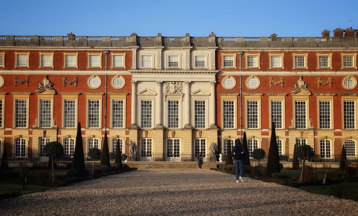 Vista ampla e iluminada pelo sol do Palácio de Hampton Court com pessoas caminhando pelo histórico caminho de cascalho, fachada simétrica e imponente e jardins formais em primeiro plano.