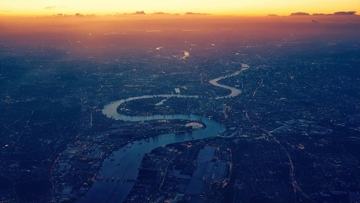 Vista aérea do rio Tâmisa serpenteando pela cidade de Londres ao pôr do sol, mostrando as curvas distintas do rio e a paisagem urbana.