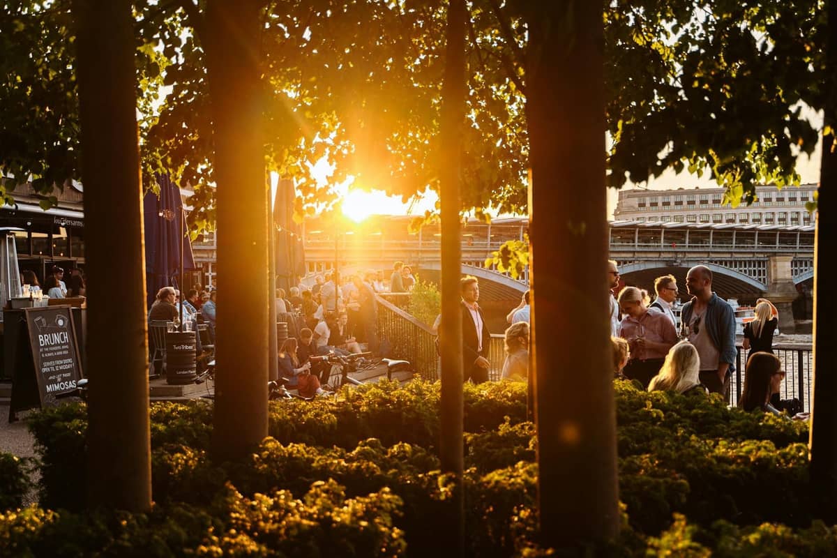 Pessoas a desfrutar de comida e bebidas em espaços ao ar livre à beira do Tamisa com pôr do sol e vegetação no centro de Londres.