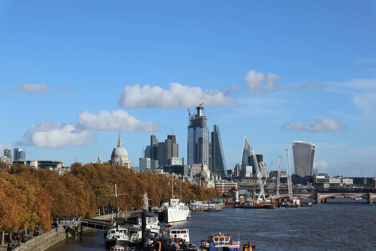 Vista do rio Tâmisa em Londres com barcos, árvores de outono e o moderno horizonte da cidade sob um céu azul limpo.