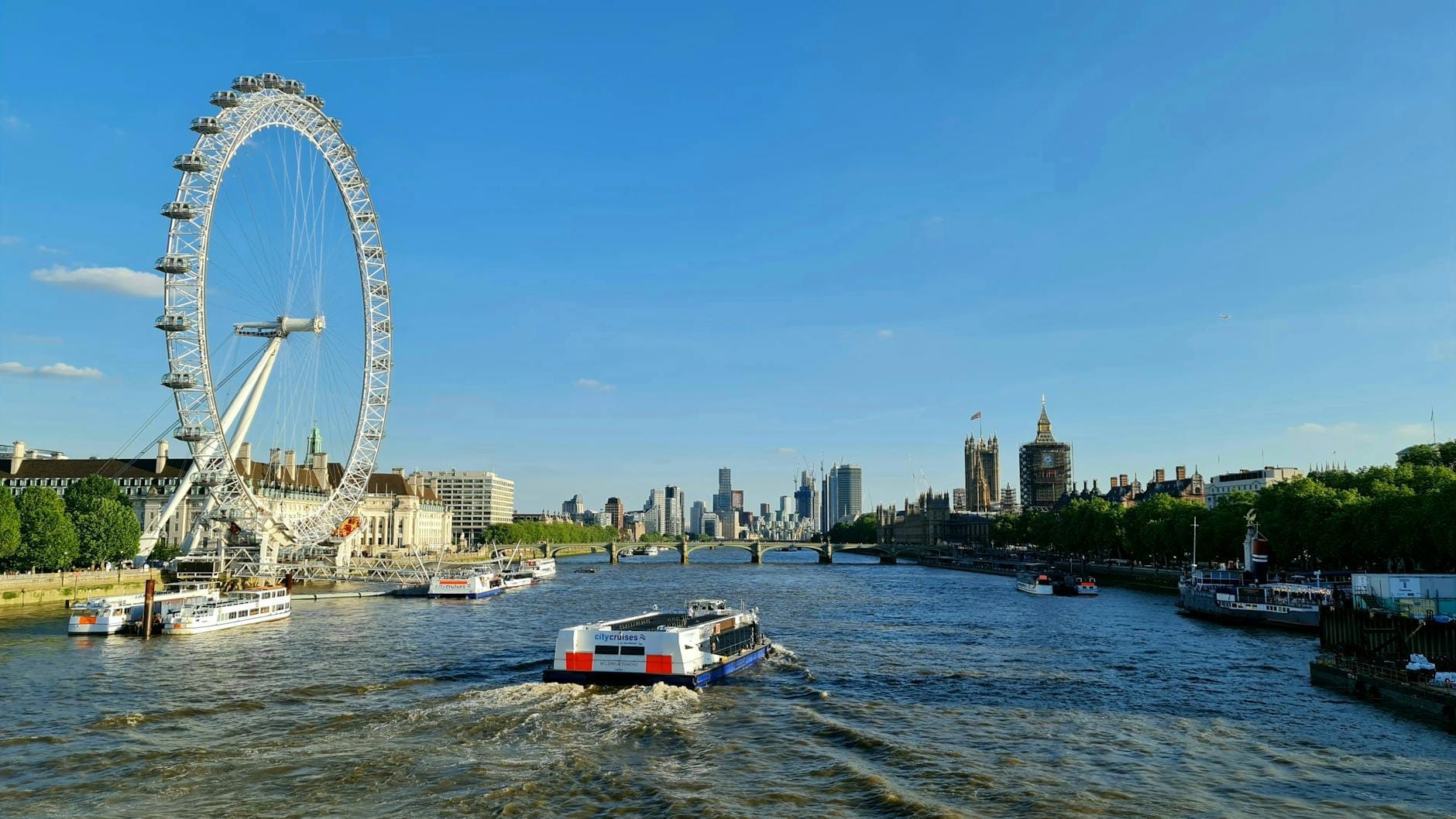 Vista do Rio Tâmisa em Londres com um barco turístico, a London Eye e famosos pontos turísticos sob um céu azul e limpo.