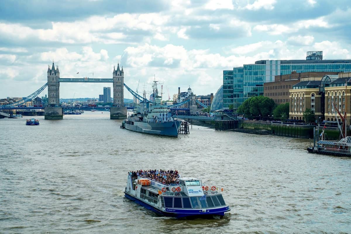 Barco de cruzeiro turístico cheio de passageiros no rio Tâmisa com a Tower Bridge e os edifícios modernos de Londres ao fundo.