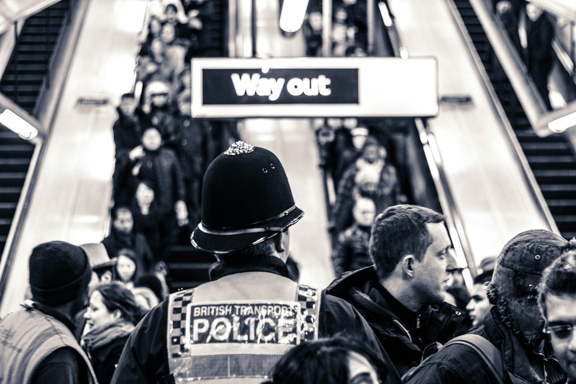 Um policial da British Transport Police está em meio a uma movimentada estação do metrô de Londres, com pessoas passando pelas escadas rolantes e uma placa de 'Saída' acima.