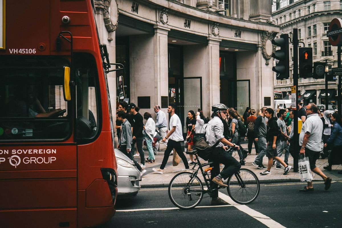 Movimentada cena de rua em Londres com ônibus vermelho, ciclista e pedestres atravessando no semáforo.