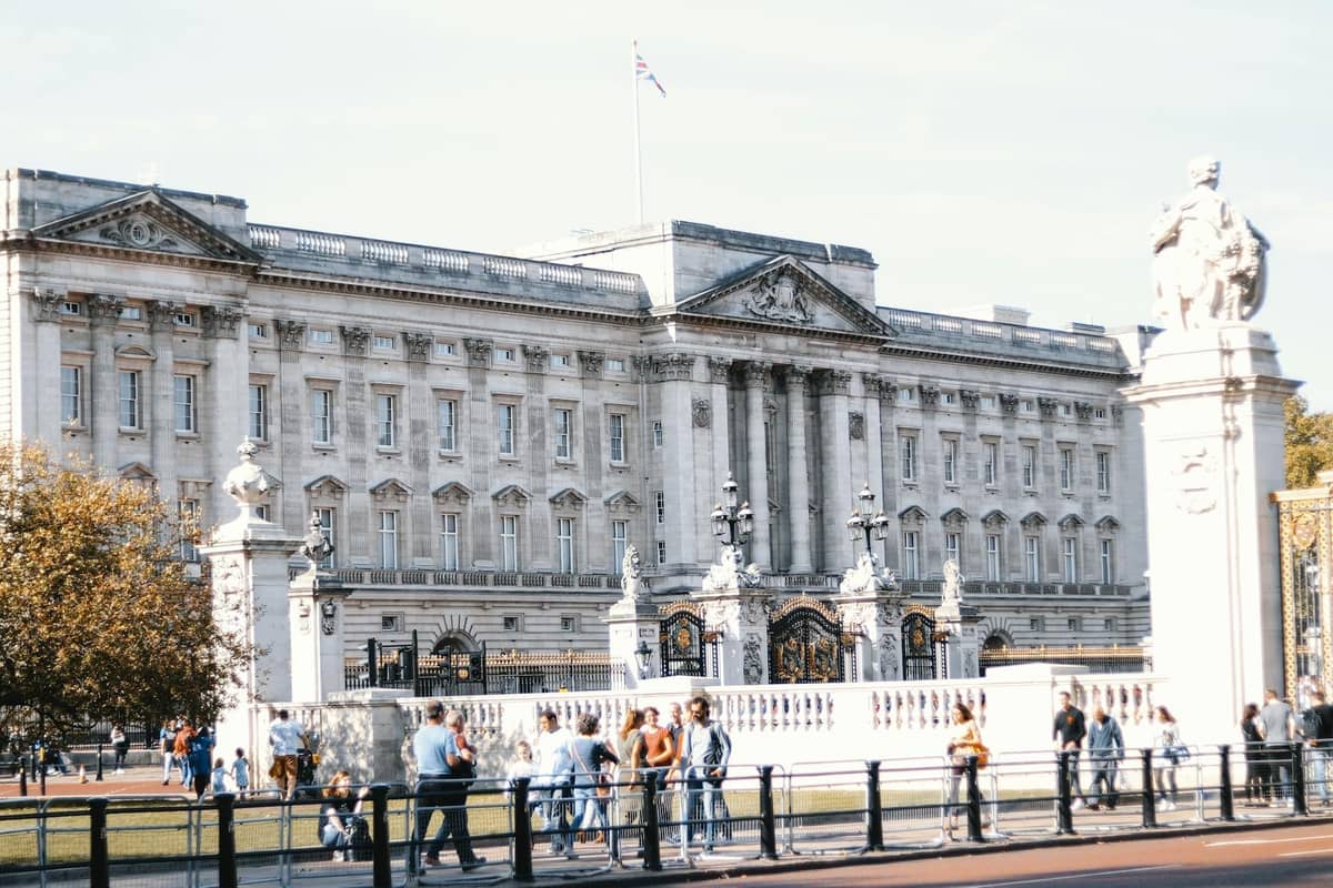 Turistas caminhando em frente ao Buckingham Palace em um dia ensolarado, com céu limpo e multidões visíveis.