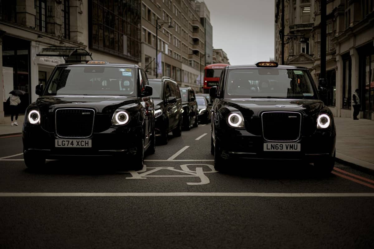 Dois icônicos táxis pretos de Londres em uma rua movimentada com outros veículos e a arquitetura clássica da cidade visível.