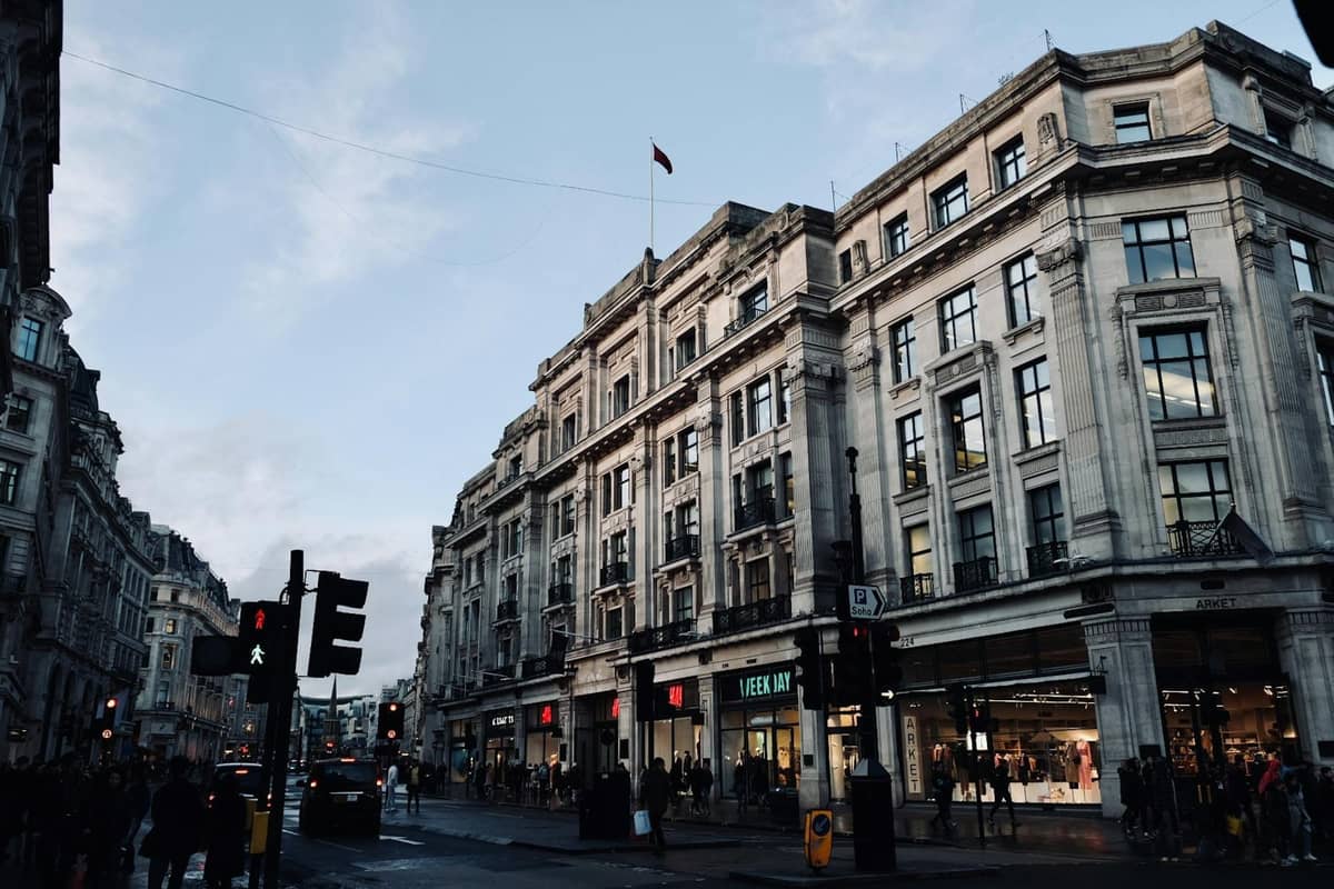 Movimentada rua comercial de Londres com grandes edifícios históricos e lojas no nível da rua, pessoas caminhando e postes de iluminação à vista.