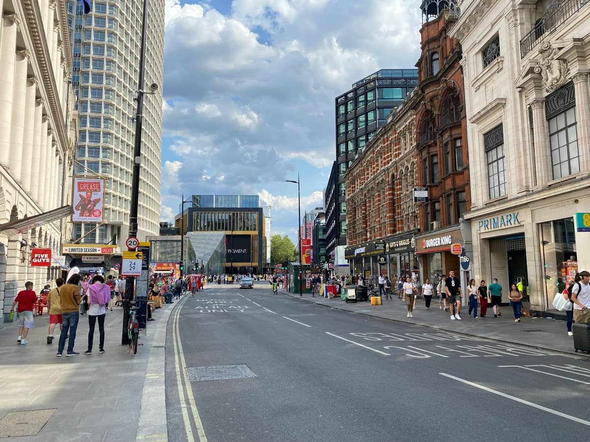 Rua movimentada de Londres com diversas lojas, compradores e arquitetura histórica sob um céu azul.