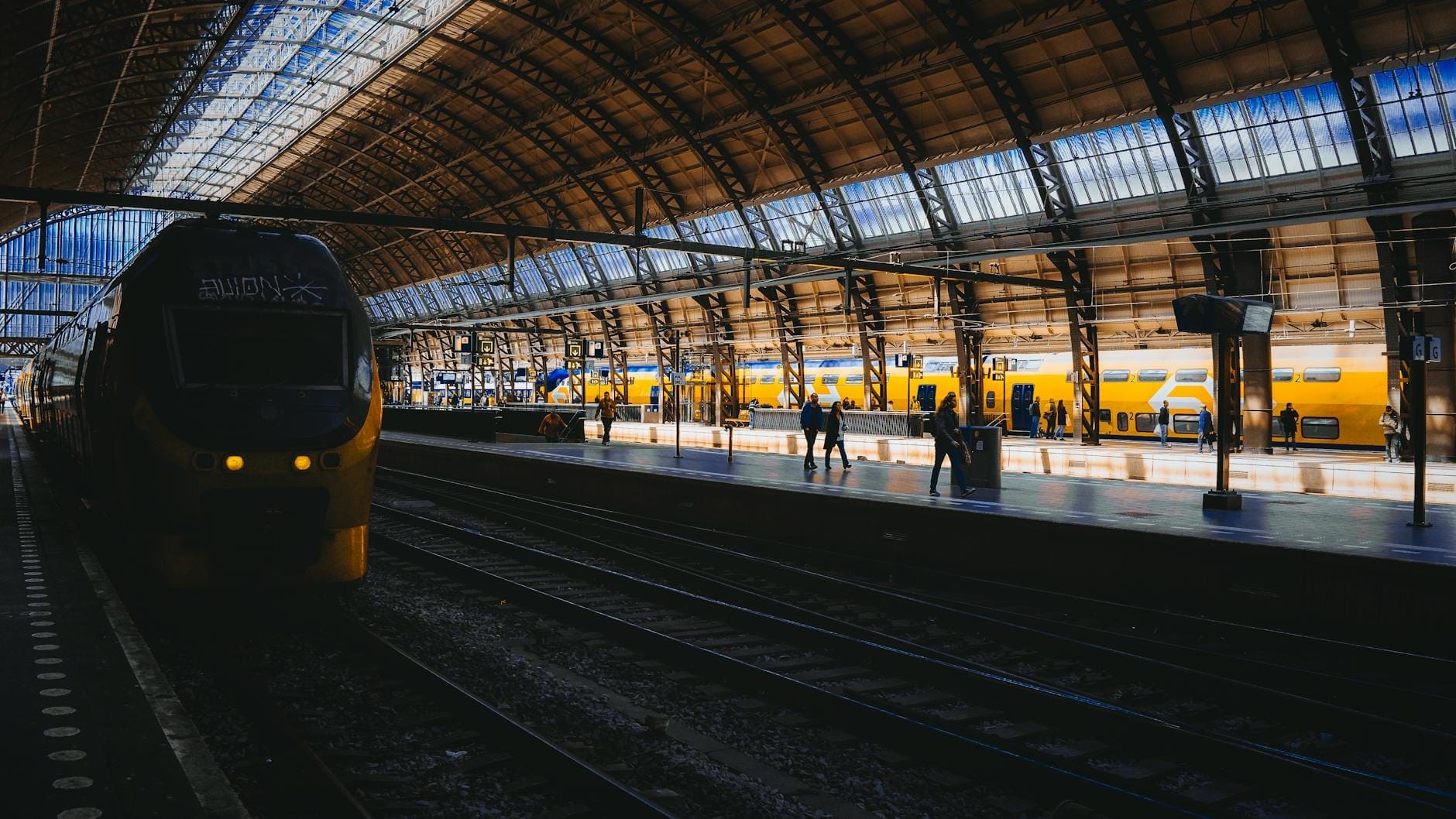 Trens amarelos modernos em uma estação ampla e bem iluminada com teto arqueado de vidro, viajantes na plataforma, evocando a jornada entre grandes cidades europeias.