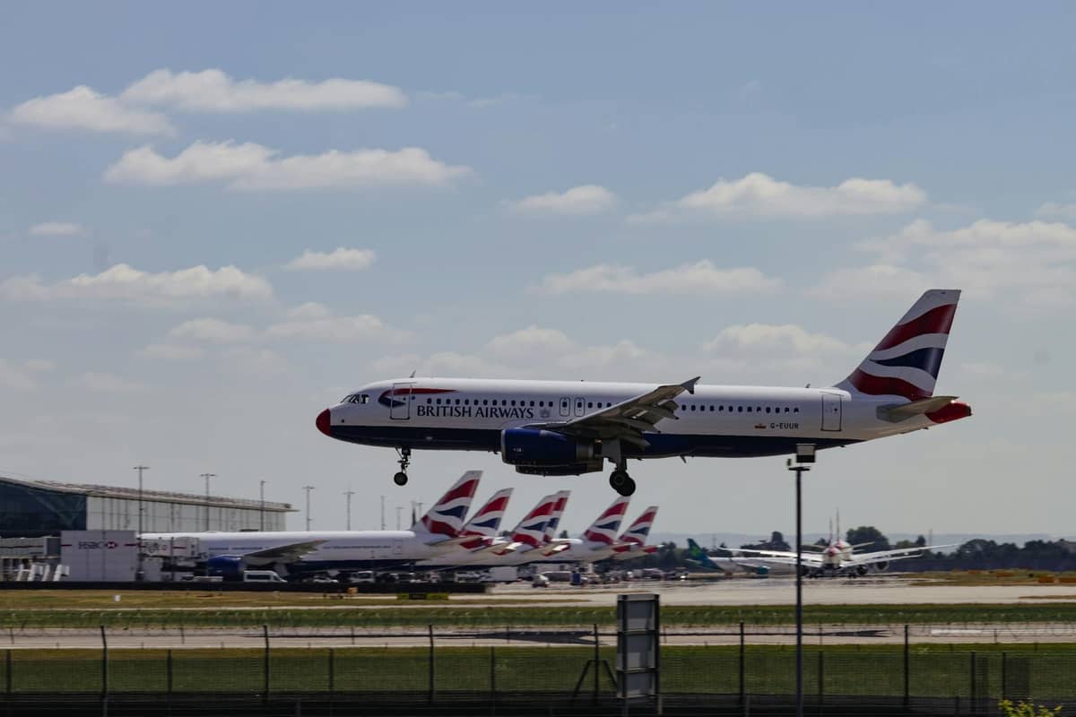 Avião da British Airways decolando em um grande aeroporto de Londres com outros aviões da British Airways e o terminal do aeroporto ao fundo