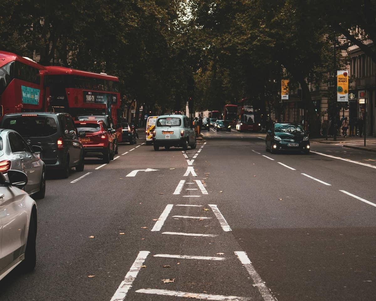 Trânsito típico de uma rua de Londres com carros, táxis pretos e ônibus vermelhos de dois andares sob árvores verdes