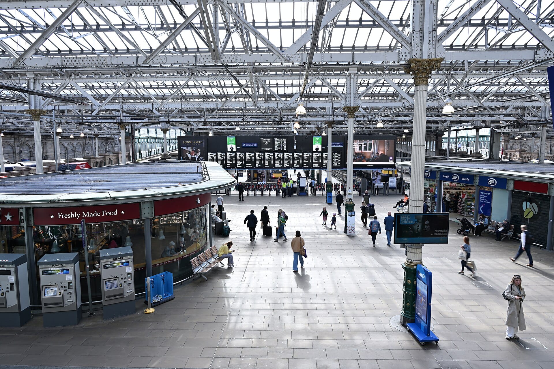 Makulay na concourse ng Edinburgh Waverley railway station na may mga pasahero, tindahan, makina ng tiket, at mga pangunahing board ng pagalis na nakikita.