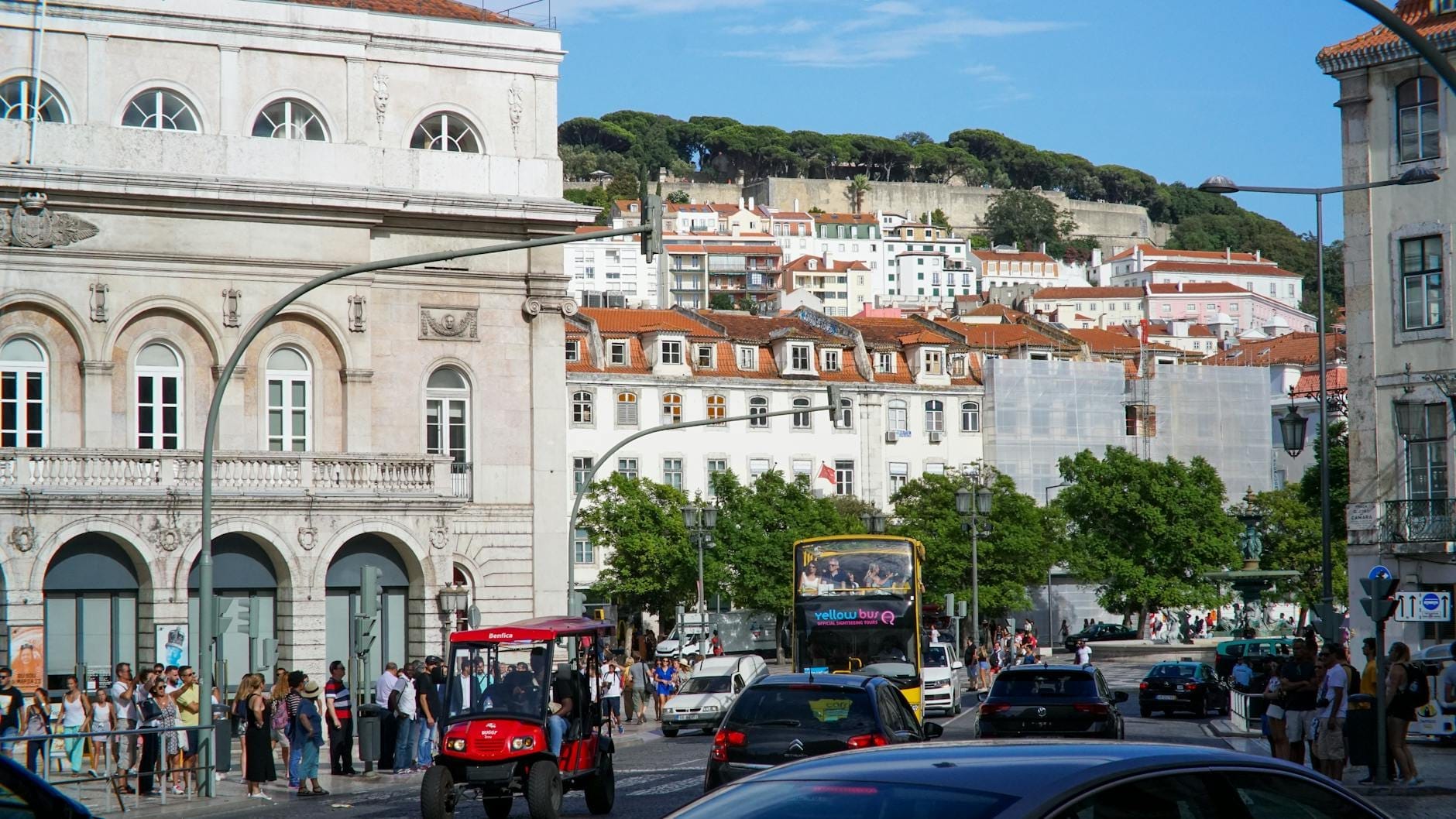 Movimentada cena de rua em Lisboa com autocarros, carros, arquitetura histórica e uma fortaleza no topo de uma colina ao fundo num dia ensolarado.