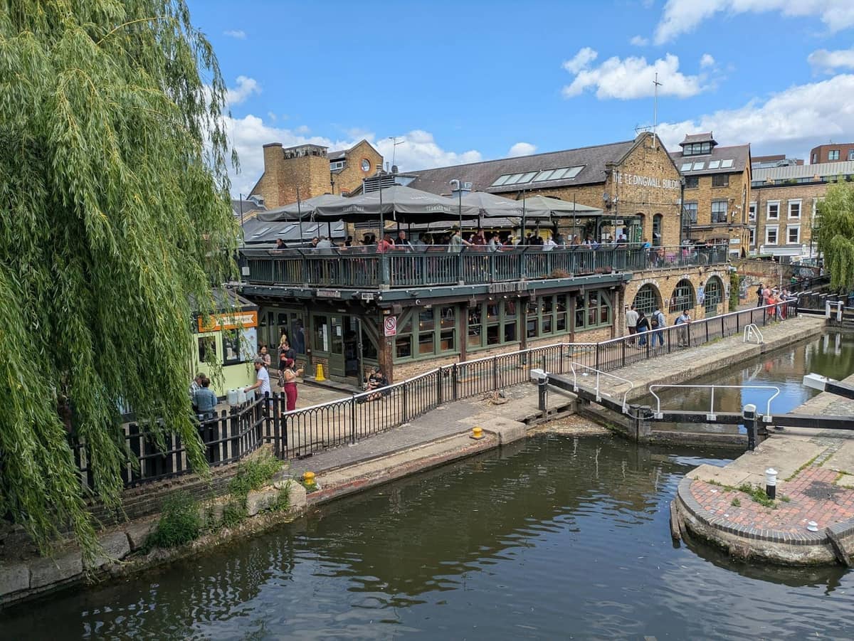 Uma animada cena à beira do canal no Camden Market em Londres, com pessoas caminhando ao lado da água, edifícios históricos e uma comporta visível sob um céu azul.