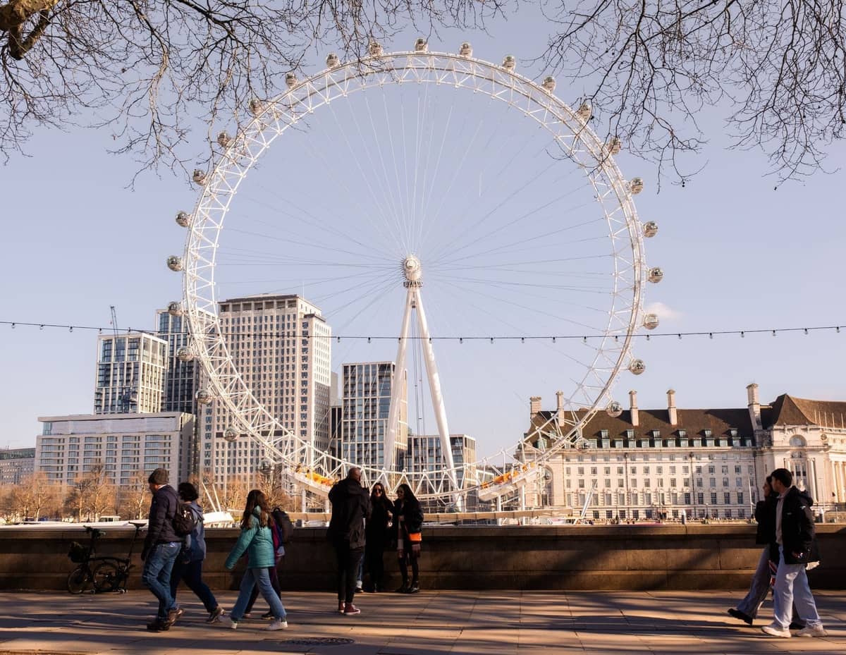 Pessoas caminhando pelo South Bank com o London Eye e edifícios da cidade ao fundo em um dia claro.