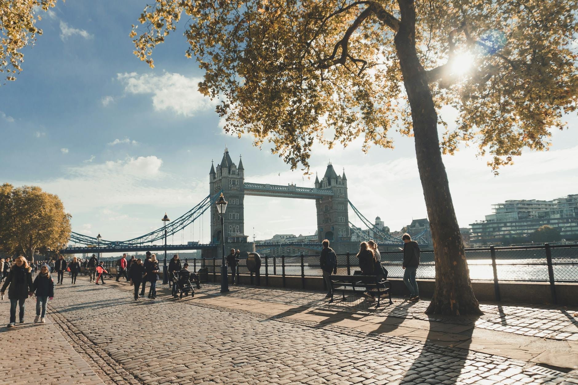 Pessoas caminhando pelo Thames Path com a Tower Bridge ao fundo em um dia ensolarado em Londres.