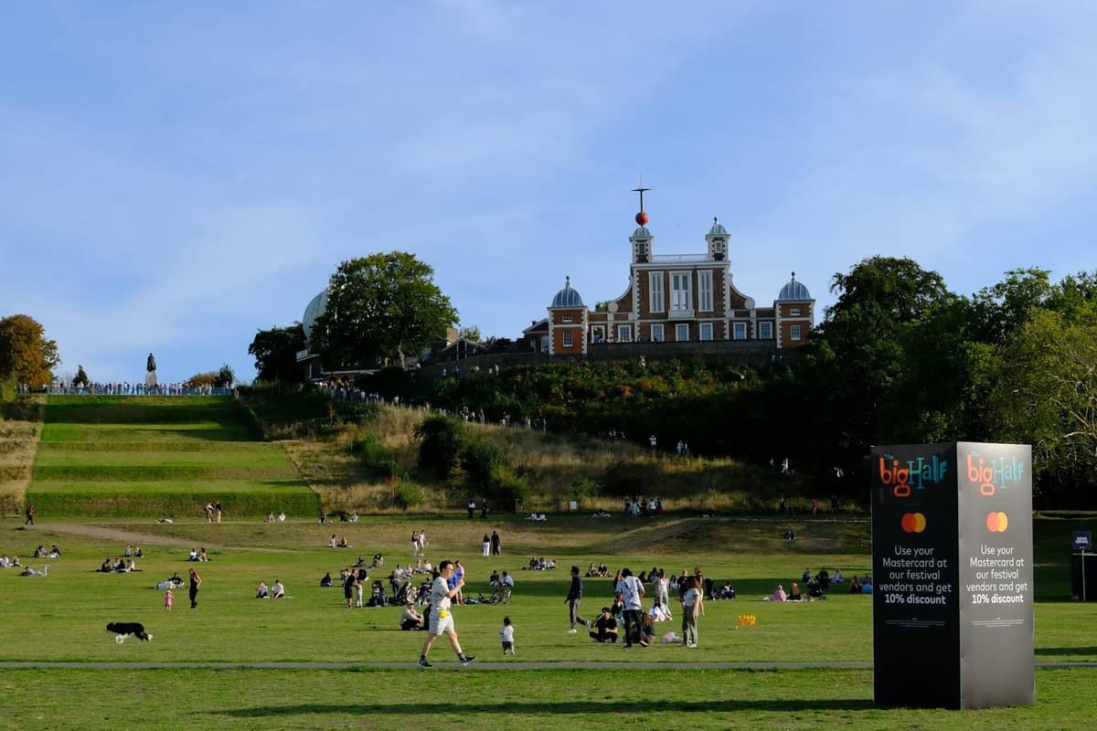 Famílias e crianças brincando em Greenwich Park com o Royal Observatory e o edifício do Meridiano Principal visíveis na colina sob um céu azul.