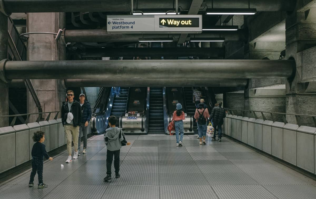 Famílias com crianças caminhando dentro de uma estação do London Underground com escadas rolantes e sinalização 'Way out' no alto.