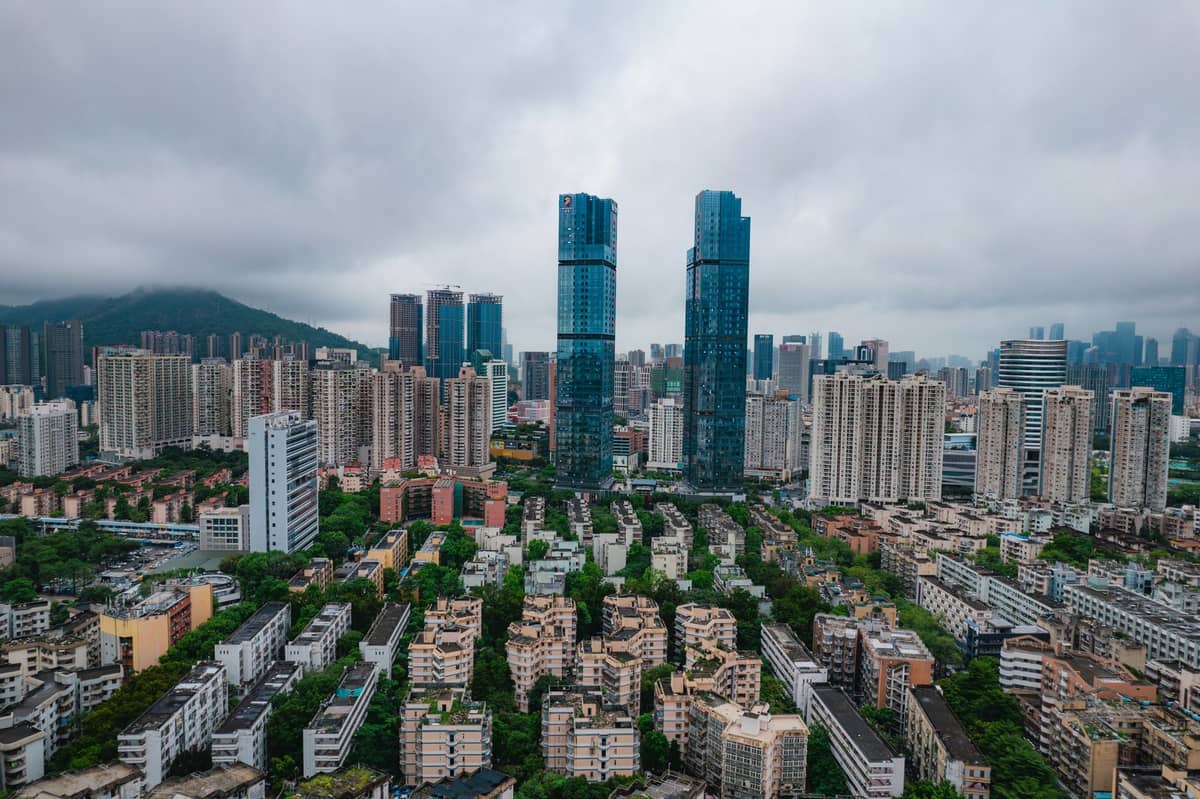 Vista aérea do Distrito de Longhua em Shenzhen com habitações densas, modernas torres e morros cobertos de nuvens ao fundo.