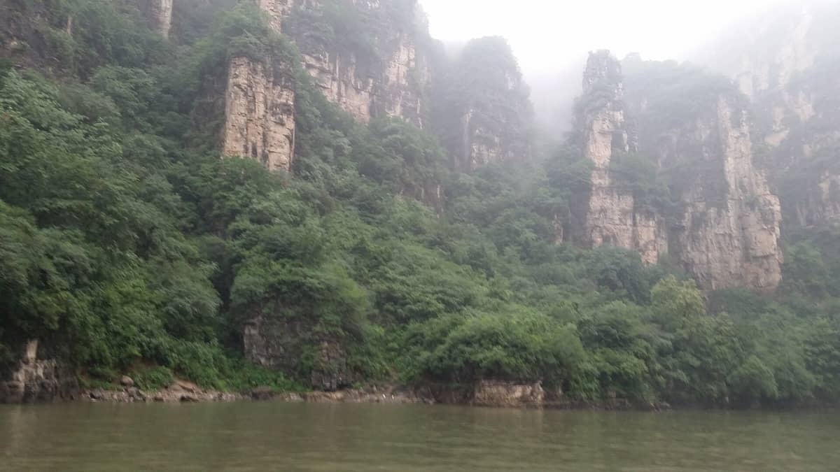 Nebelverhangene Kalksteinklippen ragen steil über dem Fluss der Longqing-Schlucht auf, die steilen Canyonwände dicht mit grüner Vegetation bewachsen.