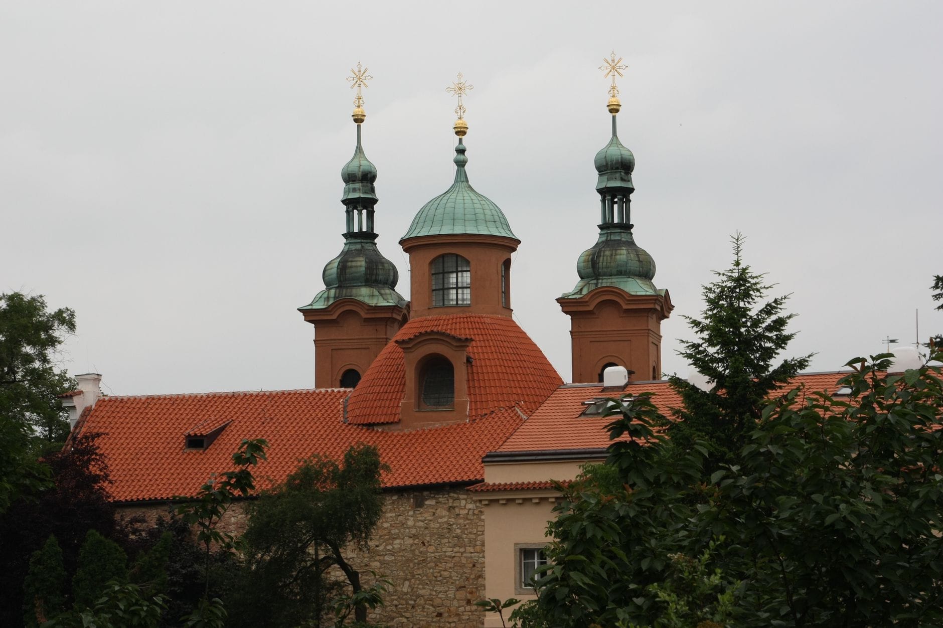 Nahaufnahme der markanten grün bekuppelten Barockfassade des Loreto-Heiligtums mit roten Ziegeldächern, eingerahmt von Bäumen im Prager Stadtteil Hradčany.