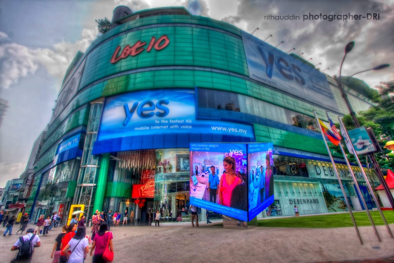 The exterior of Lot 10 Shopping Centre in Bukit Bintang features a curved glass facade, vibrant digital billboards, and crowds gathered near the main entrance.