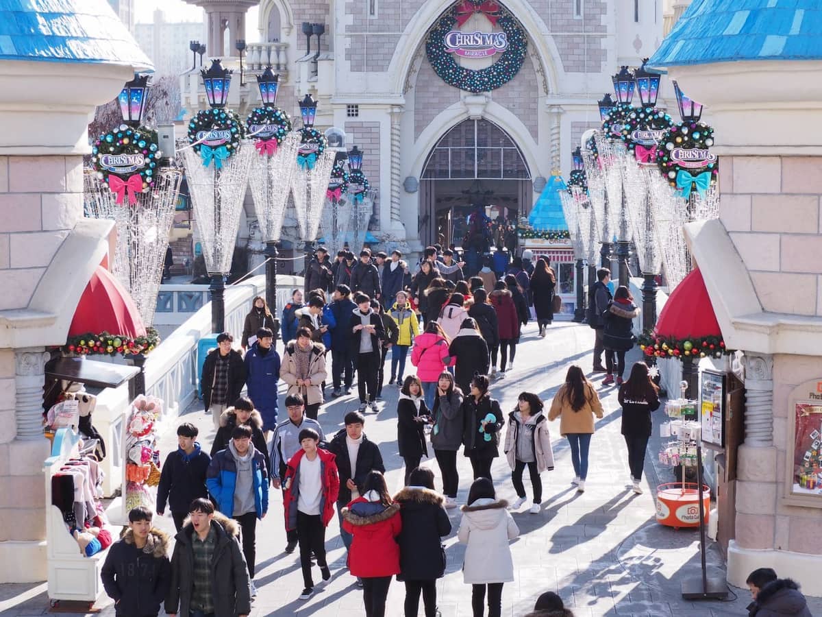 Visitantes caminham sob decorações natalinas festivas na entrada do Lotte World Adventure, cercados por telhados azuis brilhantes e luz solar de inverno.