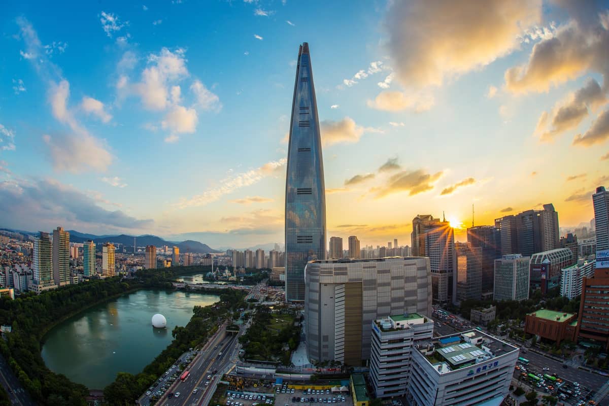 Lotte World Tower ao pôr do sol com prédios da cidade e o Lago Seokchon em primeiro plano, sob um céu amplo e colorido em Seul, Coreia do Sul.