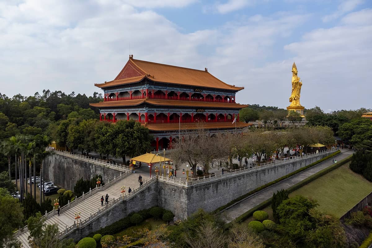 Lotus Mountain Scenic Area with traditional pavilion, large golden Guanyin statue, stone stairway, and lush greenery under a partly cloudy sky.